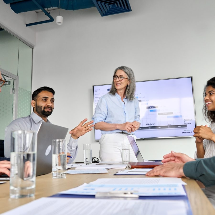 Diverse executive team people discussing company strategy at board meeting. Multicultural employees with senior leader listening indian manager brainstorming working sitting at table in office.