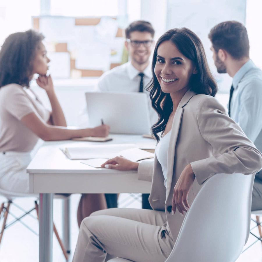 Happy to be in team. Beautiful cheerful woman looking at camera with smile while sitting at the office table with her coworkers