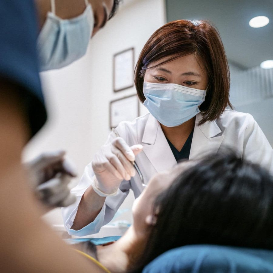 Female dentist wearing surgical mask treating woman. Male coworker is assisting mid adult healthcare worker. Medical professionals are examining patient in clinic.