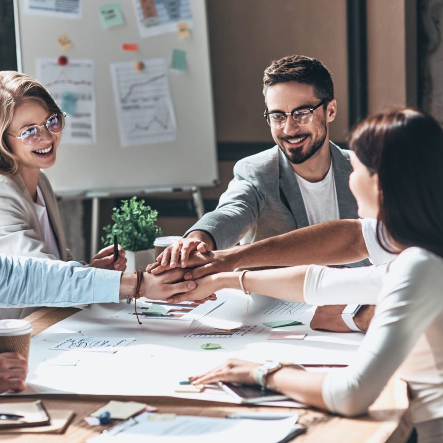 Teamwork. Young modern people in smart casual wear holding hands on top of one another in a symbol of unity while working in the creative office